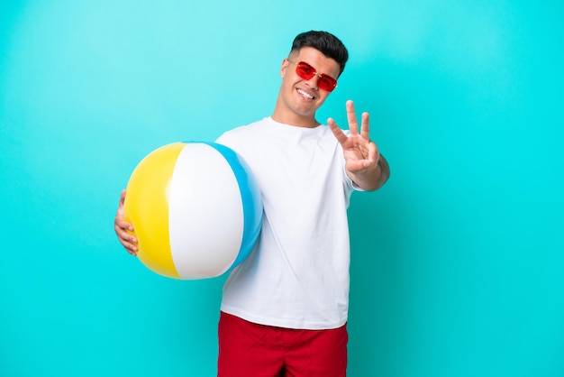 Junger kaukasischer mann, der einen wasserball lokalisiert auf blauem hintergrund hält, glücklich und drei mit den fingern zählend