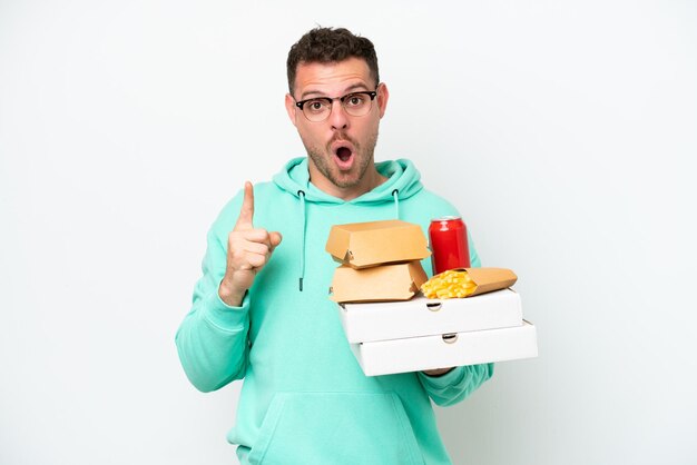 Junger kaukasischer Mann, der Fast Food isoliert auf weißem Hintergrund hält und beabsichtigt, die Lösung zu realisieren, während er einen Finger hochhebt