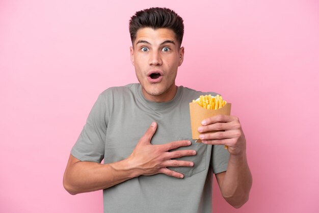 Junger kaukasischer Mann, der gebratene Chips isoliert auf rosafarbenem Hintergrund hält, überrascht und schockiert, während er nach rechts schaut