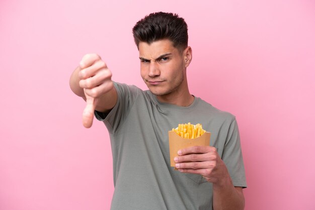 Junger kaukasischer Mann, der gebratene Chips isoliert auf rosafarbenem Hintergrund hält und den Daumen nach unten mit negativem Ausdruck zeigt