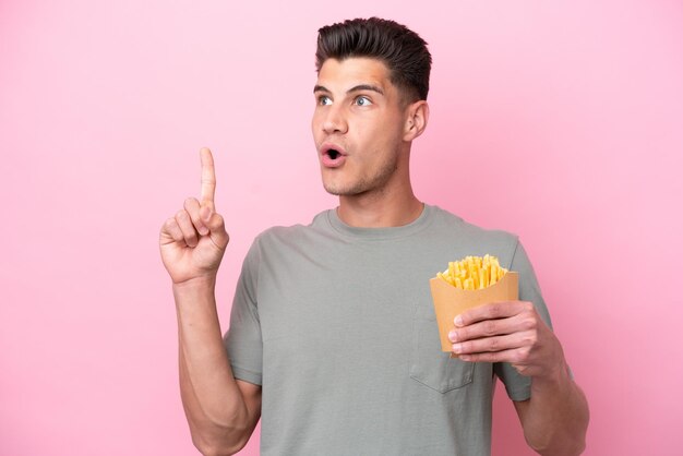 Junger kaukasischer Mann, der gebratene Chips isoliert auf rosafarbenem Hintergrund hält und eine Idee denkt, die mit dem Finger nach oben zeigt