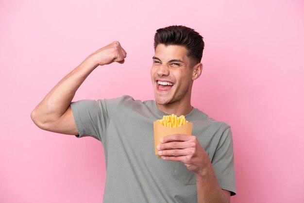 Junger kaukasischer Mann, der gebratene Chips isoliert auf rosafarbenem Hintergrund hält und einen Sieg feiert