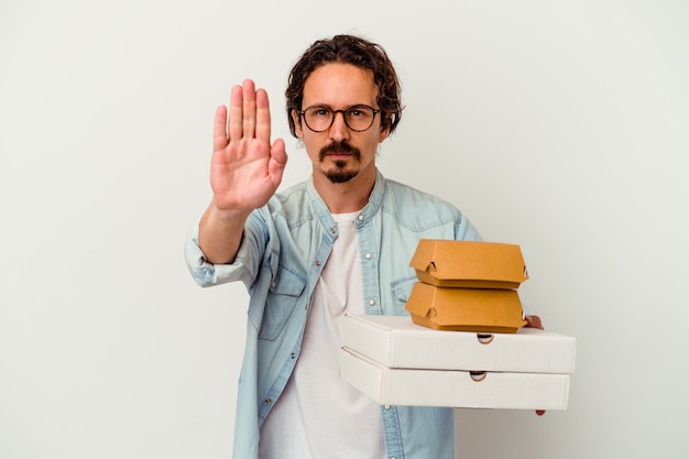 Junger kaukasischer Mann, der Hamburger eine Pizza lokalisiert auf weißer Wand hält, die mit ausgestreckter Hand steht, die Stoppschild zeigt, das Sie verhindert.