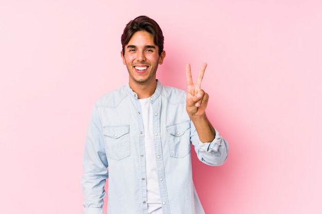 Junger kaukasischer Mann, der in einer rosa Wand posiert, die Siegeszeichen zeigt und breit lächelt.