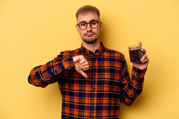 Junger kaukasischer Mann, der Kaffeedose isoliert auf gelbem Hintergrund hält und eine Abneigungsgeste zeigt, Daumen nach unten. Widerspruchskonzept.