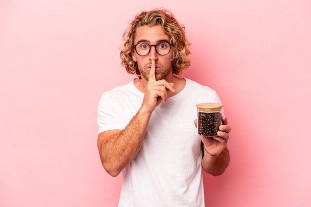 Junger kaukasischer mann, der kaffeekanne isoliert auf rosa hintergrund hält, ein geheimnis bewahrt oder um stille bittet.