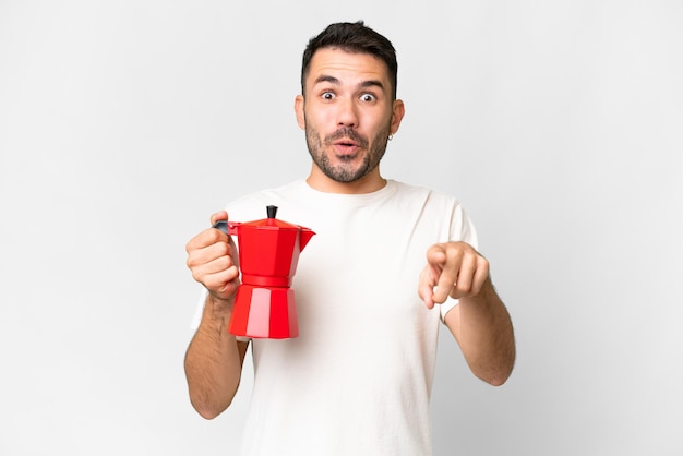 Junger kaukasischer Mann, der Kaffeekanne über isoliertem weißem Hintergrund hält, überrascht und nach vorne zeigt
