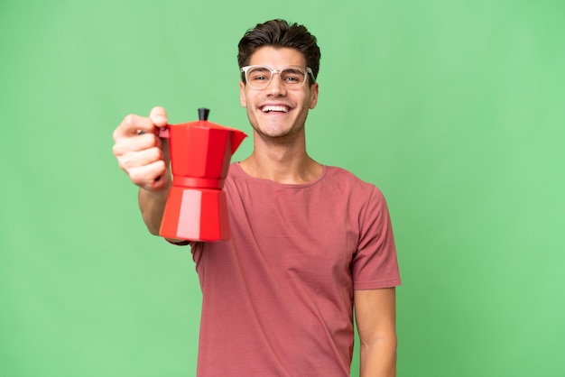 Junger kaukasischer mann, der kaffeekanne über lokalisiertem hintergrund mit glücklichem ausdruck hält