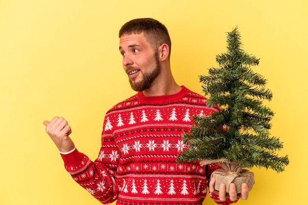 Junger kaukasischer Mann, der kleinen Baum zu Weihnachten einzeln auf gelbem Hintergrund kauft, zeigt mit Daumenfinger weg, lacht und sorglos.