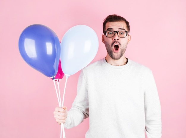 Junger kaukasischer Mann, der Luftballons mit überraschtem Ausdruck hält und einen Geburtstag feiert