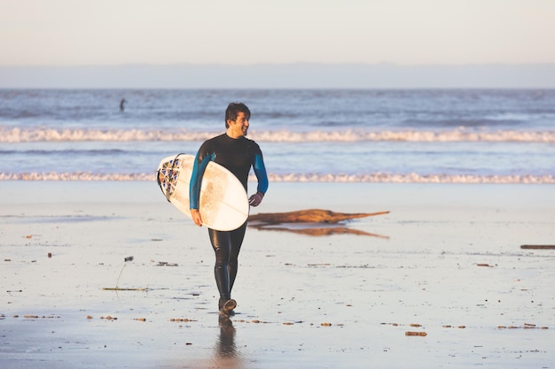 Junger kaukasischer Mann, der mit einem Surfbrett am Strand spazieren geht.