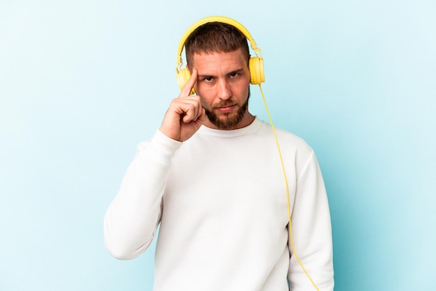 Junger kaukasischer Mann, der Musik hört, die auf blauem Hintergrund isoliert ist und mit dem Finger auf den Tempel zeigt, denkt, konzentriert sich auf eine Aufgabe.