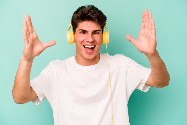 Junger kaukasischer Mann, der Musik hört, isoliert auf blauem Hintergrund, erhält eine angenehme Überraschung, aufgeregt und hebt die Hände