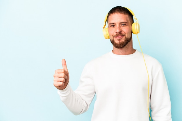 Junger kaukasischer Mann, der Musik hört, isoliert auf blauem Hintergrund, lächelt und hebt den Daumen nach oben