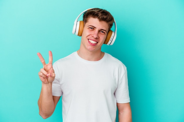 Junger kaukasischer Mann, der Musik mit Kopfhörern lokalisiert auf blauem Hintergrund freudig und sorglos zeigt, die ein Friedenssymbol mit Fingern zeigen.