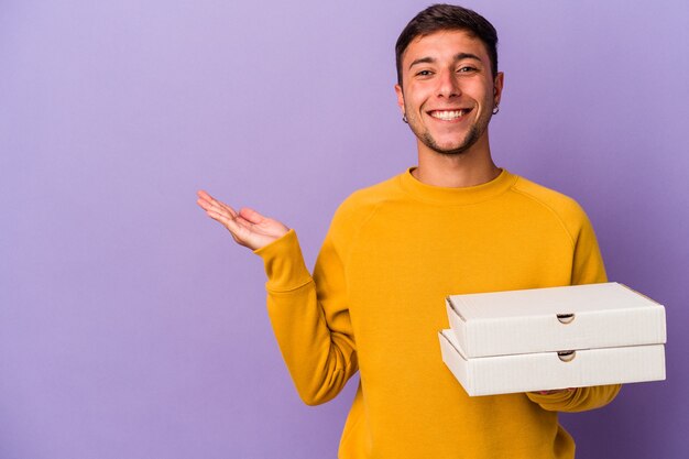 Junger kaukasischer Mann, der Pizzas einzeln auf violettem Hintergrund hält, der einen Kopienraum auf einer Handfläche zeigt und eine andere Hand an der Taille hält.