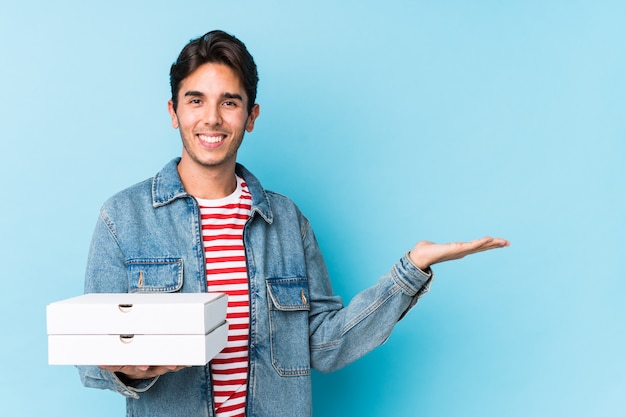 Junger kaukasischer Mann, der Pizzas lokalisiert hält, einen Kopienraum auf einer Palme zeigend und eine andere Hand auf Taille hält.