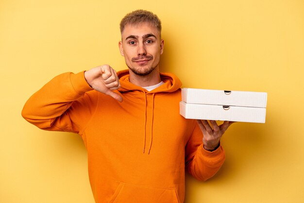 Junger kaukasischer Mann, der Pizzen isoliert auf gelbem Hintergrund hält und eine Abneigungsgeste zeigt, Daumen nach unten. Widerspruchskonzept.