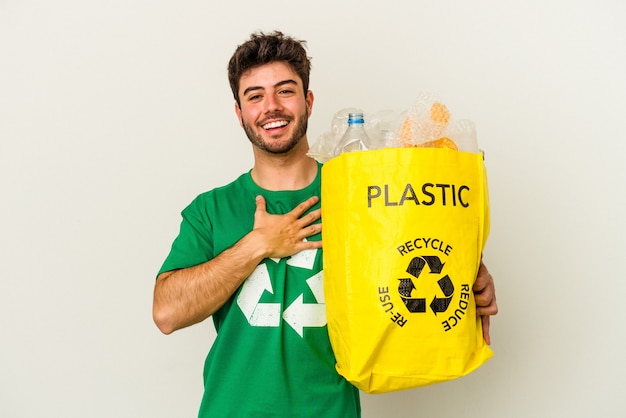 Junger kaukasischer mann, der plastik isoliert auf weißem hintergrund recycelt, lacht laut und hält die hand auf der brust.