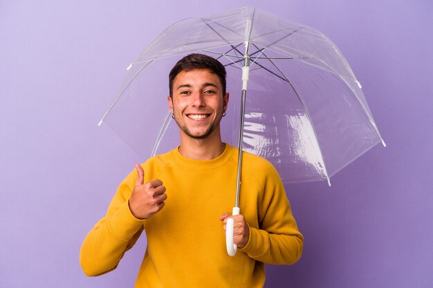 Junger kaukasischer Mann, der Regenschirm isoliert auf lila Hintergrund hält, lächelt und hebt den Daumen nach oben