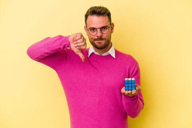 Junger kaukasischer Mann, der RubikÃ¢Â€Â™s-Würfel isoliert auf gelbem Hintergrund hält und eine Abneigungsgeste zeigt, Daumen nach unten. Widerspruchskonzept.