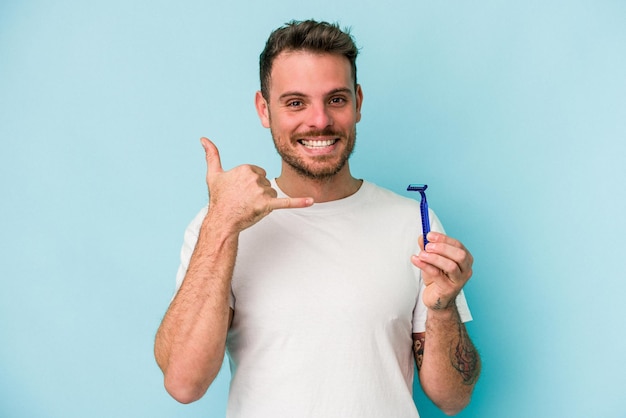 Junger kaukasischer Mann, der seinen Bart isoliert auf blauem Hintergrund rasiert und eine Handyanrufgeste mit den Fingern zeigt.