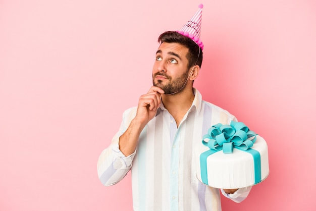 Junger kaukasischer Mann, der seinen Geburtstag lokalisiert auf rosa Wand feiert, die seitwärts mit zweifelhaftem und skeptischem Ausdruck schaut.