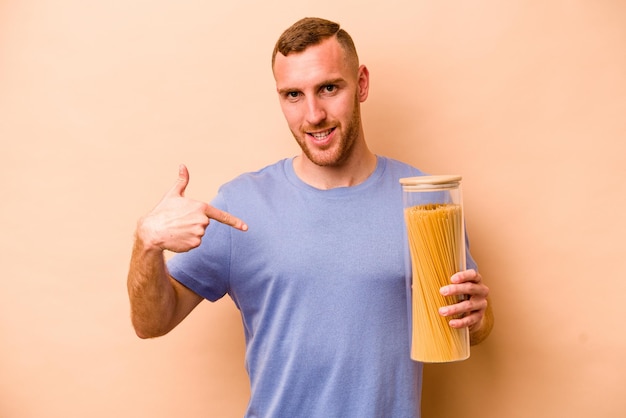 Junger kaukasischer mann, der spaghettiglas lokalisiert auf beiger hintergrundperson hält, die mit der hand auf einen hemdkopienraum zeigt, stolz und selbstbewusst