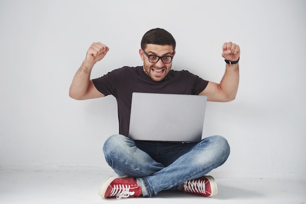 Junger kaukasischer Mann, der über weißer Backsteinmauer unter Verwendung des Computerlaptops sitzt, der in Schock mit einem Überraschungsgesicht glücklich ist, ängstlich und aufgeregt mit Angstausdruck.