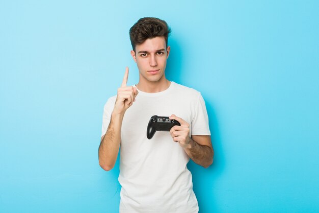 Junger kaukasischer Mann, der Videospiele mit dem Gamecontroller zeigt Nummer Eins mit dem Finger spielt.