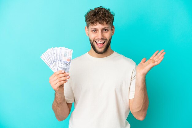 Junger kaukasischer Mann, der viel Geld auf blauem Hintergrund mit schockiertem Gesichtsausdruck nimmt