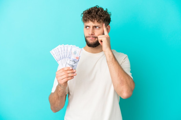 Junger kaukasischer mann, der viel geld nimmt, isoliert auf blauem hintergrund, der zweifel hat und nachdenkt