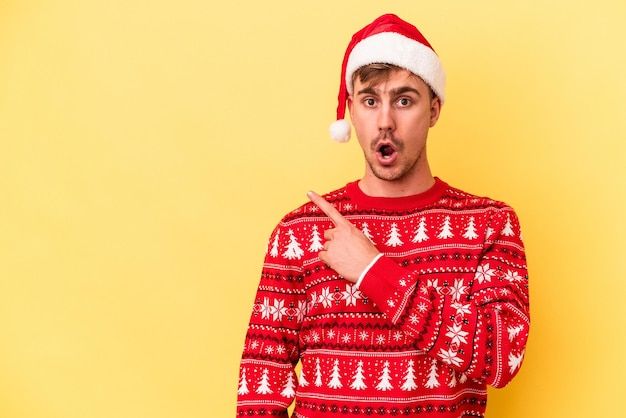 Junger kaukasischer Mann, der Weihnachten isoliert auf gelbem Hintergrund feiert, der auf die Seite zeigt