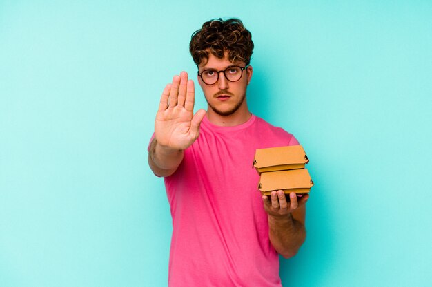 Junger kaukasischer Mann, der zwei Burger hält, die auf der blauen Wand lokalisiert stehen, die mit ausgestreckter Hand steht, die Stoppschild zeigt, das Sie verhindert.