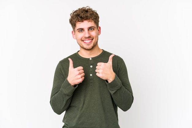 Junger kaukasischer Mann des blonden lockigen Haares, der beide Daumen anhebt, lächelnd und zuversichtlich.