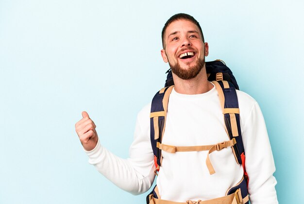 Junger kaukasischer Mann des Wanderers lokalisiert auf blauem Hintergrund zeigt mit Daumenfinger weg, lachend und sorglos.