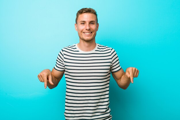 Junger kaukasischer Mann gegen eine blaue Wand zeigt unten mit den Fingern, positives Gefühl.