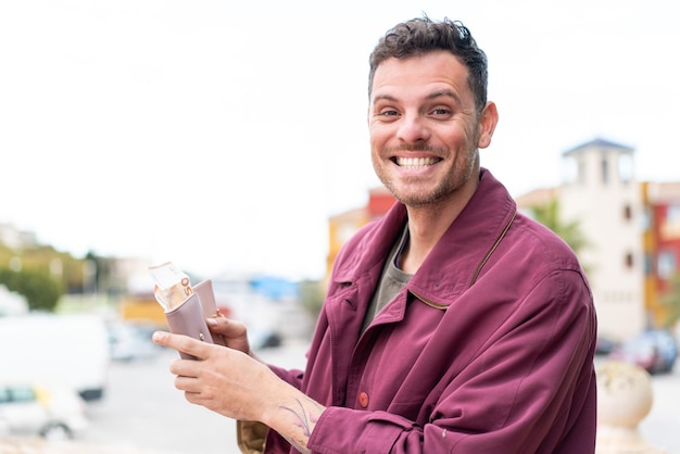 Junger kaukasischer mann im freien, der brieftasche mit geld mit glücklichem ausdruck hält