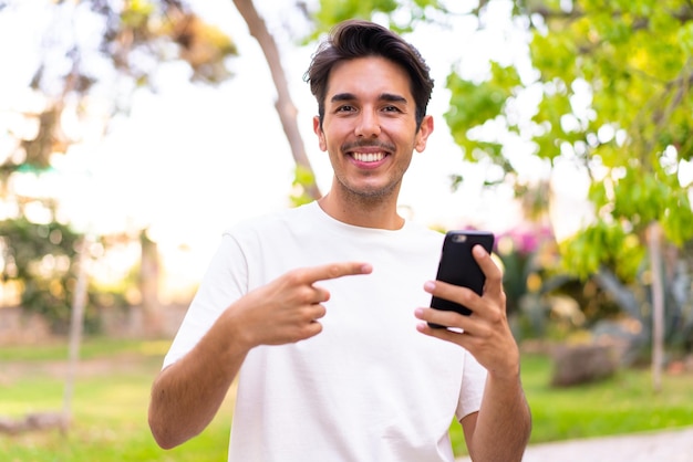 Junger kaukasischer Mann in einem Park, der Handy benutzt und darauf zeigt