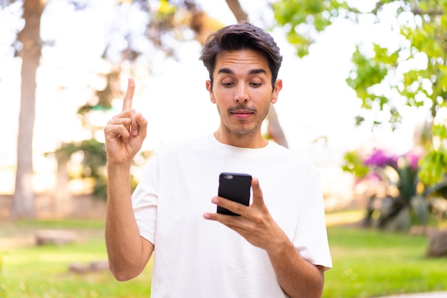 Junger kaukasischer Mann in einem Park mit Handy und hebendem Finger