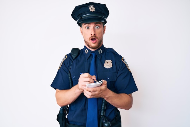 Junger kaukasischer mann in polizeiuniform schreibt fein in schockgesicht und sieht skeptisch und sarkastisch überrascht mit offenem mund aus