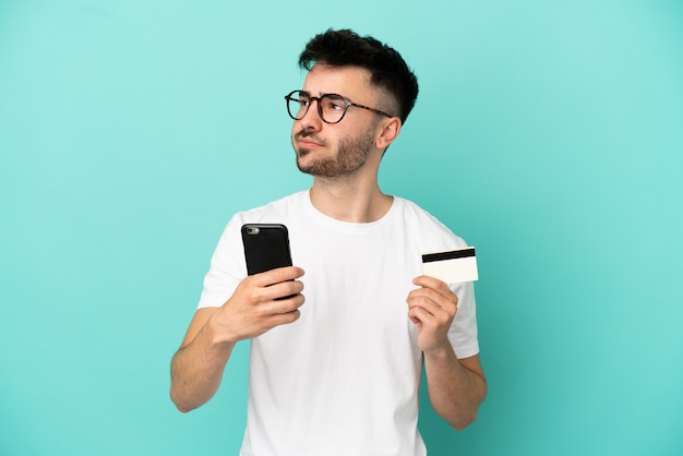 Junger kaukasischer Mann isoliert auf blauem Hintergrund, der beim Nachdenken mit dem Handy mit einer Kreditkarte kauft