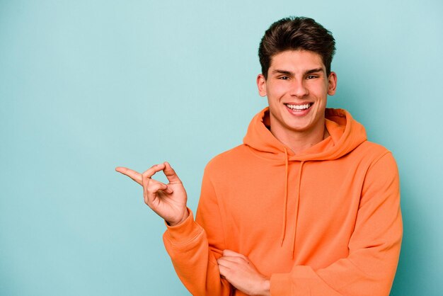 Junger kaukasischer Mann isoliert auf blauem Hintergrund, der fröhlich lächelt und mit dem Zeigefinger wegzeigt