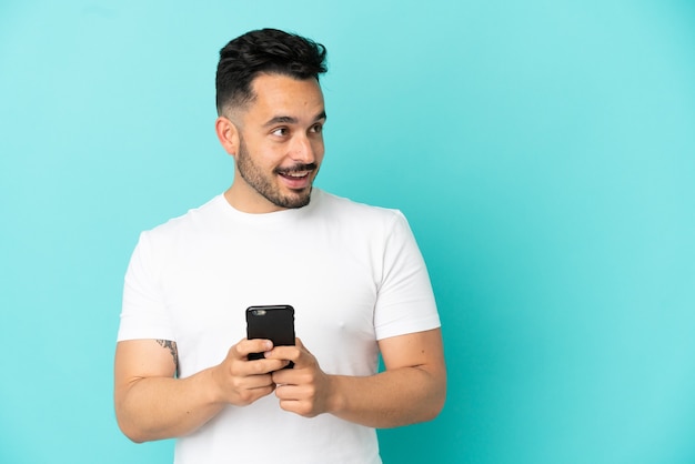 Junger kaukasischer Mann isoliert auf blauem Hintergrund, der Handy benutzt und nach oben schaut