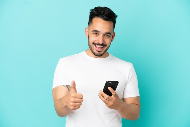 Junger kaukasischer Mann isoliert auf blauem Hintergrund mit Handy beim Daumen hoch