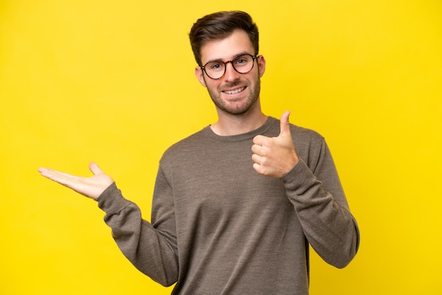 Junger kaukasischer Mann isoliert auf gelbem Hintergrund, der ein imaginäres Exemplar auf der Handfläche hält, um eine Anzeige einzufügen, und mit Daumen nach oben