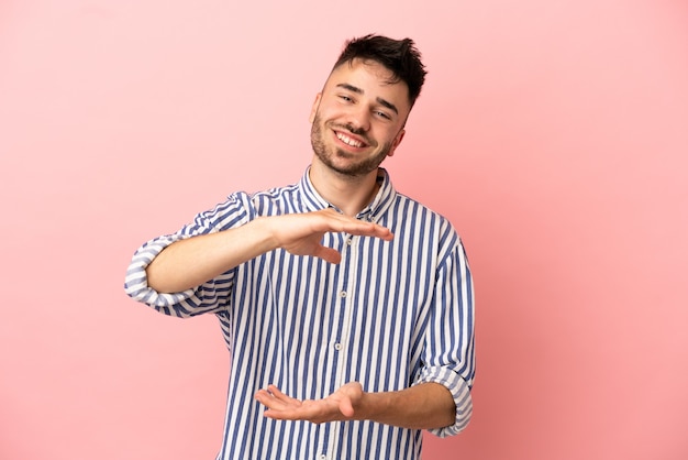 Junger kaukasischer Mann isoliert auf rosafarbenem Hintergrund, der ein imaginäres Exemplar auf der Handfläche hält, um eine Anzeige einzufügen