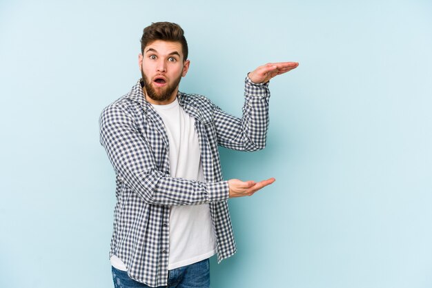 Junger kaukasischer Mann lokalisiert auf blauem Hintergrund schockiert und erstaunt, einen Kopierraum zwischen Händen haltend.