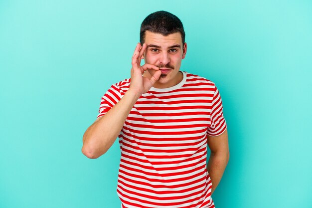 Junger kaukasischer Mann lokalisiert auf blauer Wand mit den Fingern auf den Lippen, die ein Geheimnis halten.