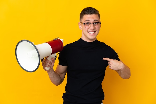 Junger kaukasischer Mann lokalisiert auf gelber Wand, die ein Megaphon und mit überraschendem Gesichtsausdruck hält